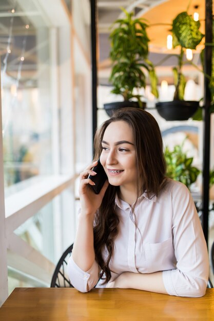 Mujer encantadora joven que llama con teléfono celular mientras está sentado solo en la cafetería durante el tiempo libre, mujer atractiva con linda sonrisa que tiene una conversación hablando con el teléfono móvil mientras descansa en la cafetería