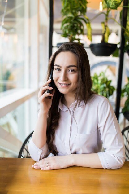 Mujer encantadora joven que llama con teléfono celular mientras está sentado solo en la cafetería durante el tiempo libre, mujer atractiva con linda sonrisa que tiene una conversación hablando con el teléfono móvil mientras descansa en la cafetería