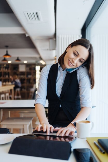 Mujer encantadora joven que llama con el teléfono inteligente mientras está sentado solo en la cafetería, conversando con el teléfono móvil