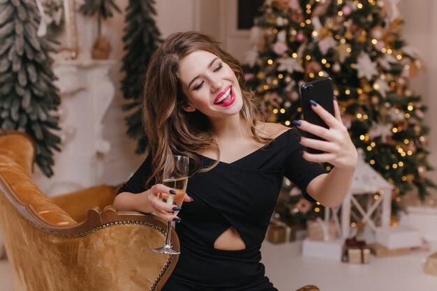 Mujer encantadora con manicura de moda haciendo selfie con champán cerca del árbol de Navidad. Retrato de interior de lindo modelo femenino blanco en traje de fiesta tomando una foto de sí misma en vacaciones.