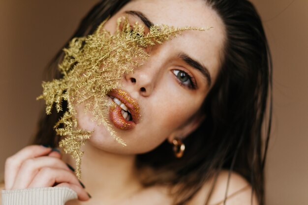 Mujer encantadora con maquillaje brillante con planta verde. Foto de primer plano de una chica morena alegre con hojas.