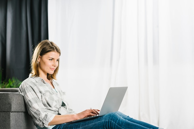 Mujer encantadora que usa la computadora portátil cerca del sofá