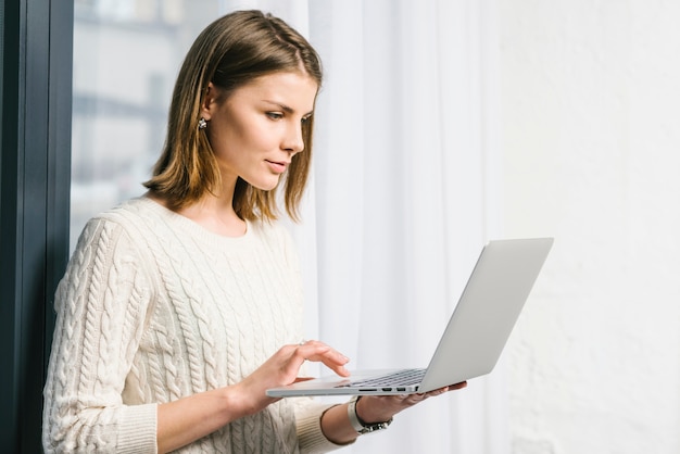 Mujer encantadora que usa la computadora portátil cerca de la ventana