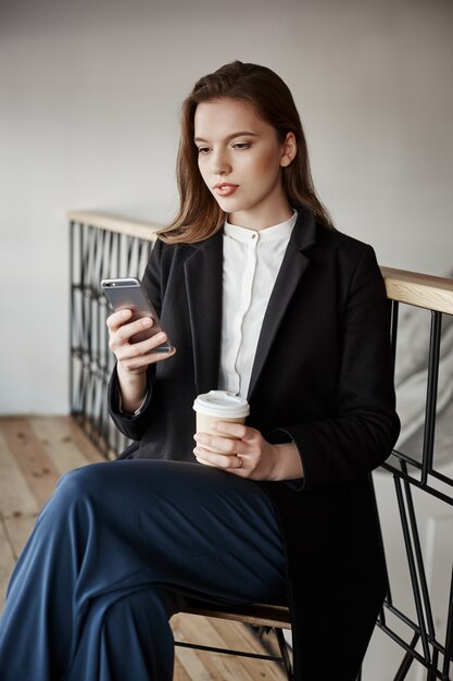 mujer encantadora en ropa elegante sentado en la cafetería, tomando café mientras sostiene el teléfono inteligente