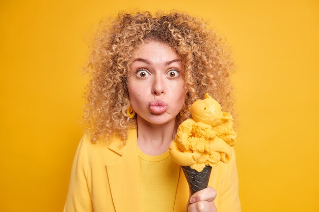 Mujer encantadora sorprendida sostiene un gran helado en gofre negro mantiene los labios doblados siente la tentación de comer un postre frío de verano vestido con ropa de moda aislada sobre una pared amarilla