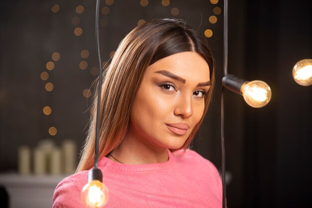 Una mujer encantadora en suéter posando junto a lámparas de luz.