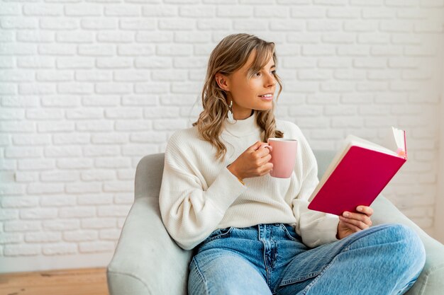 Mujer encantadora en suéter suave cálido disfrutando de una taza de té y un libro de lectura en la acogedora mañana de invierno