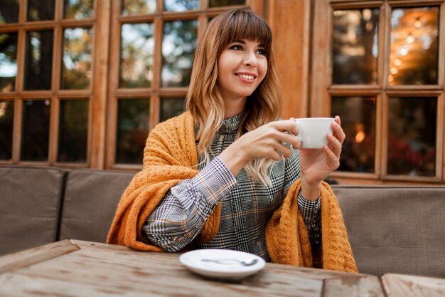 Mujer encantadora tomar un café en la acogedora cafetería con interior de madera, hablando por teléfono móvil. Sosteniendo una taza de capuchino caliente. Temporada de invierno. Con vestido elegante y cuadros amarillos.