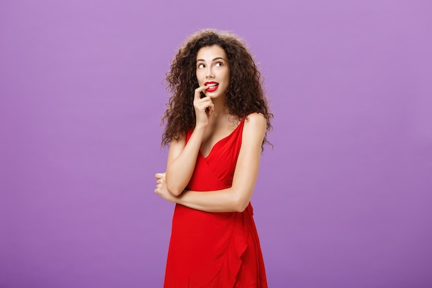 Mujer encantadora tonta y tímida con cabello oscuro y rizado en elegante vestido de noche con lápiz labial rojo mordiendo el dedo mirando lindo en la esquina superior derecha imagen pensativa o soñando con algo deseable.