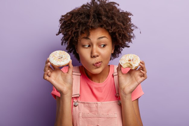 Mujer encantadora vacilante con peinado afro, frunce los labios y mira deliciosas donas glaseadas dulces que contienen muchas calorías, vestida con ropa casual, aislada sobre la pared púrpura del estudio. Comida