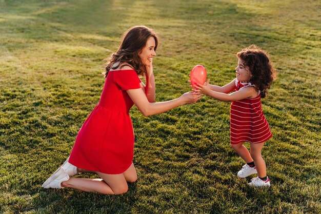 Mujer encantadora en vestido rojo jugando con su hija en el parque. Foto al aire libre de la señorita riendo mirando a la hermana pequeña con una sonrisa.