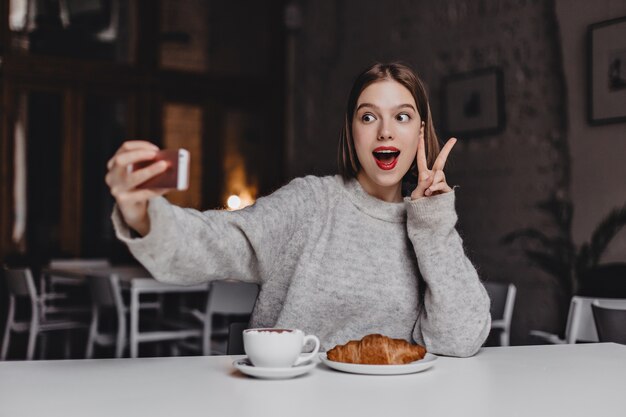 Mujer enérgica en suéter gris y lápiz labial rojo hace selfie. Retrato de niña mostrando el signo de la paz en la cafetería con croissant en la mesa.