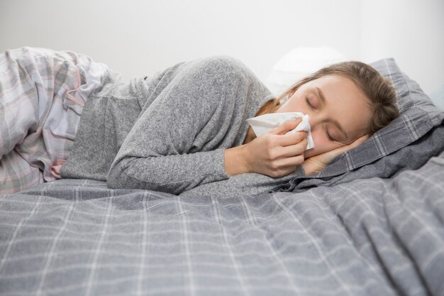 Mujer enferma acostada en la cama con los ojos cerrados, soplando la nariz