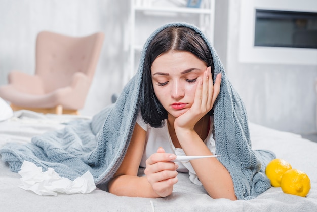 Mujer enferma acostada en la cama sufriendo de fiebre mirando termómetro