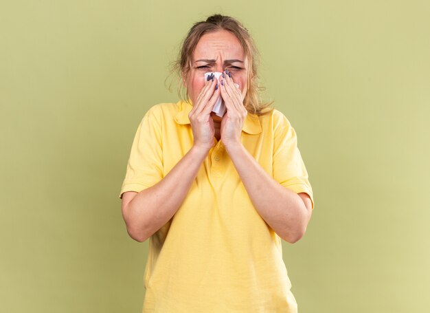 Mujer enferma en camisa amarilla que se siente terrible que sufre de gripe y frío que sopla la nariz estornudando en el tejido de pie sobre la pared verde