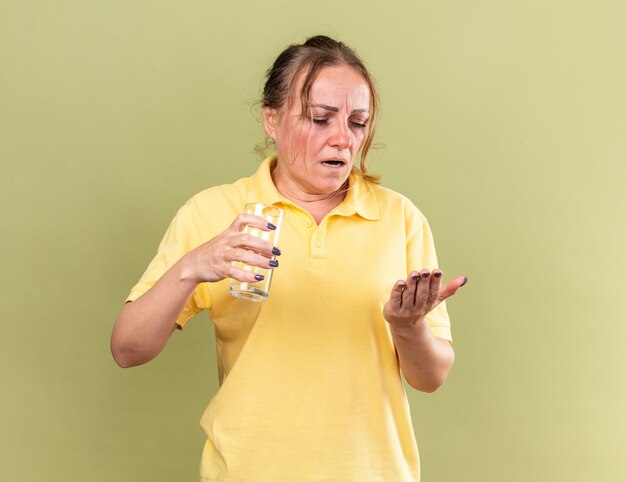 Mujer enferma en camisa amarilla que se siente terrible sosteniendo un vaso de agua y pastillas para tomar medicamentos que sufren de gripe y resfriado de pie sobre una pared verde