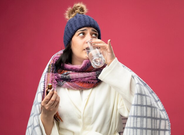 Mujer enferma caucásica joven débil vestida con bata, gorro y bufanda de invierno, vaso de agua mezclada con medicamento