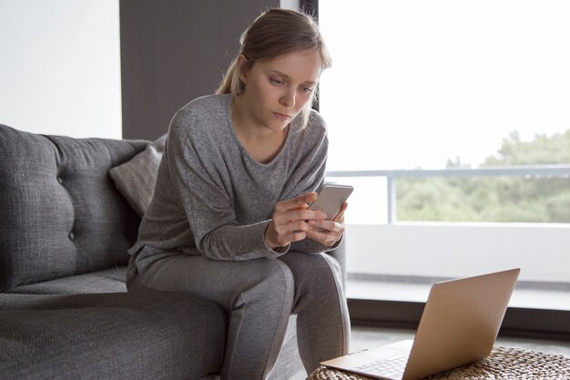 Mujer enferma concentrada sentada en casa, enviando mensajes de texto al teléfono