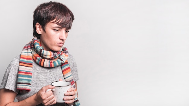 Mujer enferma contempló con la bufanda multicolora alrededor de su cuello que sostiene la taza contra fondo gris