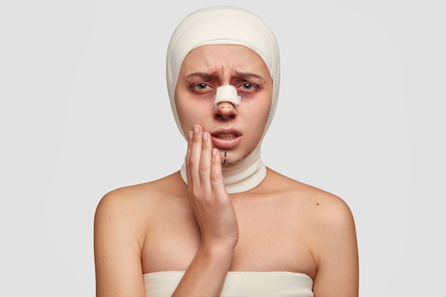Una mujer enferma desesperada mantiene la mano en la mejilla, sufre un fuerte dolor después de un injerto de piel, tiene un vendaje o yeso en la nariz