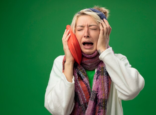 Foto gratuita mujer enferma enferma molesta con pelo corto en bufanda caliente y sombrero sintiéndose mal sosteniendo una botella de agua para mantenerse caliente llorando frustrado sufrimiento de gripe de pie sobre fondo verde