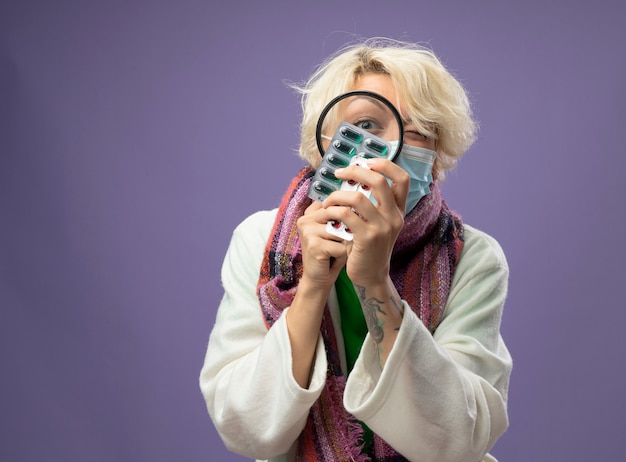 Foto gratuita mujer enferma enferma con pelo corto en bufanda caliente y máscara protectora facial mirando pastillas a través de una lupa de pie sobre una pared púrpura