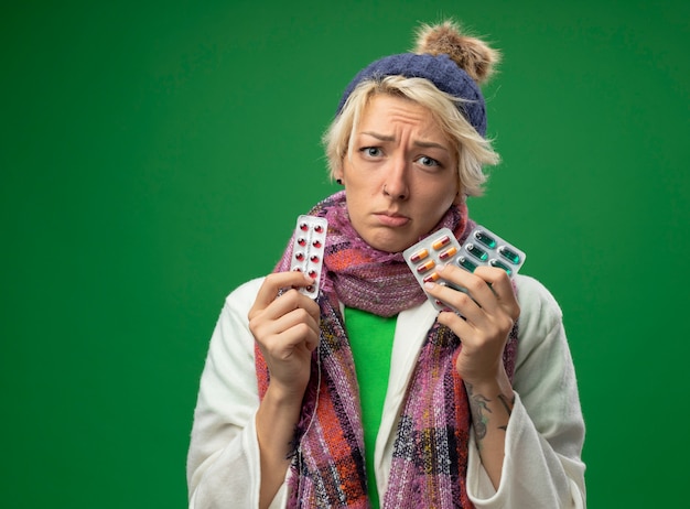 Foto gratuita mujer enferma enferma con el pelo corto en bufanda caliente y sombrero que se siente mal sosteniendo ampollas con pastillas que está molesto e infeliz de pie sobre la pared verde