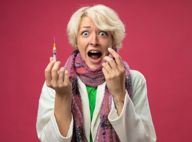 Mujer enferma enferma con el pelo corto en la bufanda que se siente mal sosteniendo una ampolla y una jeringa gritando de pánico de pie sobre una pared rosa