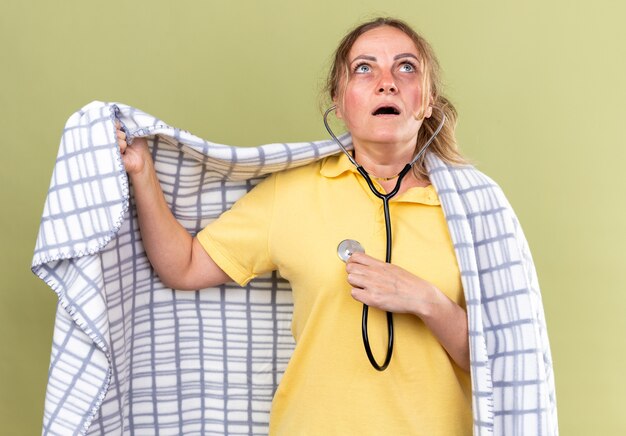 Mujer enferma envuelta en una manta que se siente mal que sufre de gripe y frío escuchando los latidos de su corazón con un estetoscopio que parece preocupada de pie sobre una pared verde