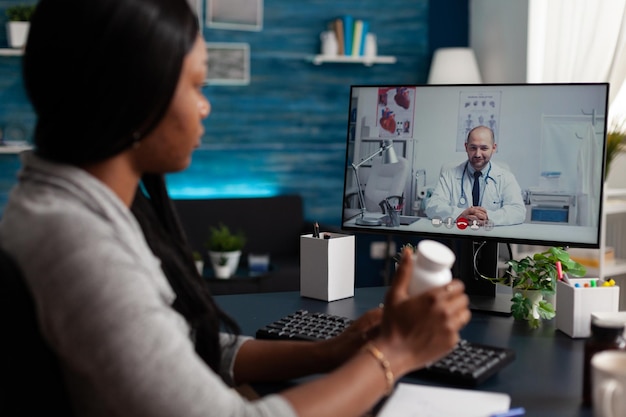 Mujer enferma con frasco de píldoras en videollamada con el médico, haciendo consulta remota en la computadora. Paciente preguntando al médico sobre el tratamiento sanitario en una videoconferencia en línea en casa.