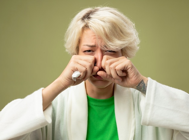 Mujer enferma insalubre con cabello corto que se siente mal por estar molesto frotándose los ojos que sufren de goteo nasal de pie sobre fondo claro