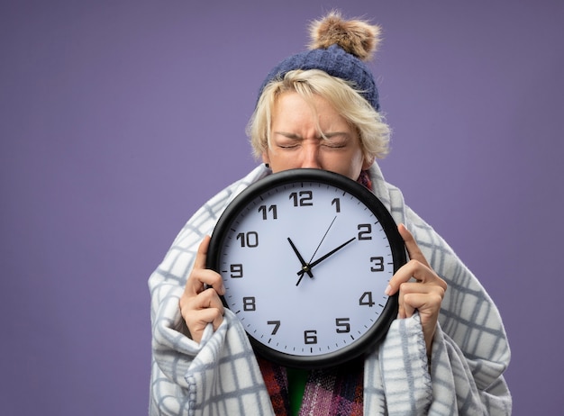 Mujer enferma insalubre con el pelo corto con bufanda caliente y gorro sintiéndose mal envuelto en una manta con reloj de pared con los ojos cerrados scaredover pared púrpura