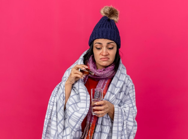Mujer enferma joven concentrada con gorro de invierno y bufanda envuelta en cuadros añadiendo medicamento en un vaso de agua aislado en la pared rosa con espacio de copia