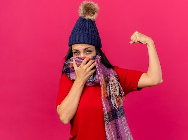 Mujer enferma joven confiada con gorro de invierno y bufanda cubriendo la boca con bufanda mirando al frente haciendo un gesto fuerte aislado en la pared rosa con espacio de copia