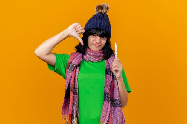 Mujer enferma joven disgustada con gorro de invierno y bufanda sosteniendo el termómetro mirando al frente mostrando el pulgar hacia abajo aislado en la pared naranja con espacio de copia