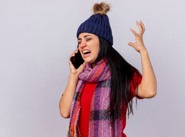 Mujer enferma joven enojada con gorro de invierno y bufanda de pie en la vista de perfil hablando por teléfono manteniendo la mano en el aire con los ojos cerrados aislados en la pared blanca