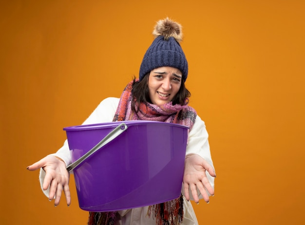 Foto gratuita mujer enferma joven irritada con gorro de invierno bata y bufanda con náuseas estirando un cubo de plástico hacia el frente mirando al frente aislado en la pared naranja