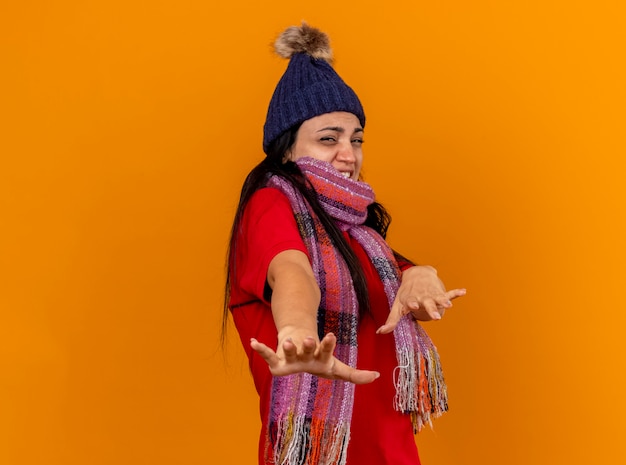 Mujer enferma joven molesta con gorro de invierno y bufanda de pie en la vista de perfil estirando las manos hacia el frente mirando al frente gesticulando no aislado en la pared naranja