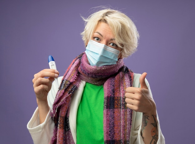 Mujer enferma malsana con el pelo corto en bufanda caliente y máscara protectora facial sosteniendo termómetro mostrando los pulgares para arriba sonriendo de pie sobre la pared púrpura