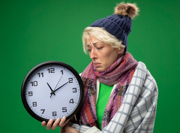Foto gratuito mujer enferma malsana con el pelo corto en bufanda caliente y sombrero sintiéndose mal envuelto en una manta con reloj de pared mirándolo con expresión triste sobre fondo verde