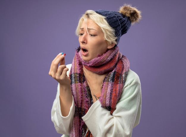 Foto gratuita mujer enferma malsana con el pelo corto en bufanda caliente y sombrero sintiéndose mal sosteniendo pastillas mirándolos preocupado de pie sobre fondo púrpura