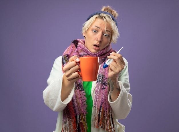 Mujer enferma no saludable con pelo corto en bufanda caliente y sombrero sintiéndose mal sosteniendo una taza de té caliente y un termómetro mirando la taza confundida y preocupada de pie sobre fondo púrpura