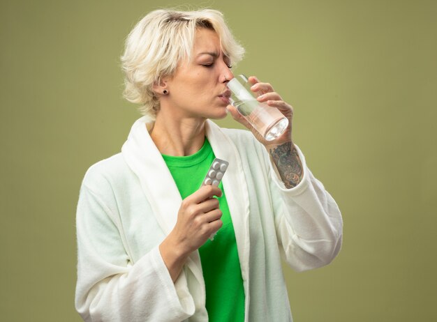 Mujer enferma con el pelo corto sintiéndose mal sosteniendo un vaso de agua tomando medicamentos de pie sobre una pared de luz