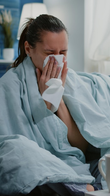 Mujer enferma que usa tejido para sonarse la nariz que moquea y tiene frío. Adulto infeliz que se siente enfermo y bebe té de una taza y se sienta en el sofá con una manta. Persona con tratamiento de enfermedades y medicamentos.