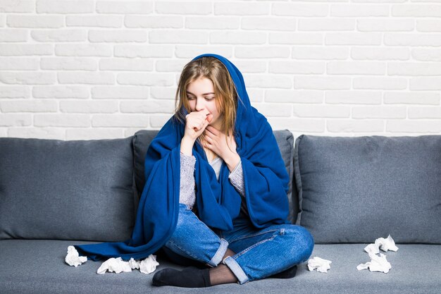 Mujer enferma con reum y dolor de cabeza sosteniendo una servilleta, sentada en el sofá con colcha y pastillas en casa