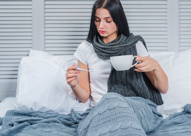 Mujer enferma sosteniendo una taza de café mirando el termómetro