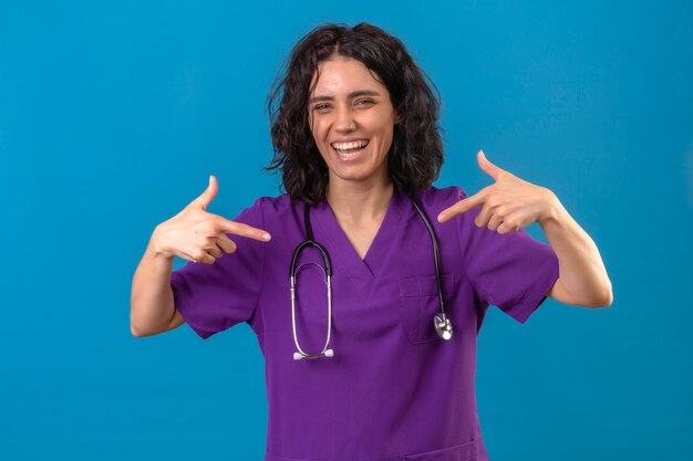 Mujer enfermera en uniforme médico y un estetoscopio apuntando a sí misma con los pulgares mirando alegre sonriendo alegremente en azul aislado