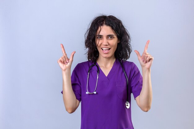 Mujer enfermera en uniforme médico y con estetoscopio con cara feliz sonriendo señalando con el dedo hacia arriba de pie en blanco aislado