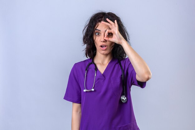 Mujer enfermera en uniforme médico y con estetoscopio divirtiéndose haciendo bien firmar con los dedos mirando a través de este cartel de pie en blanco aislado