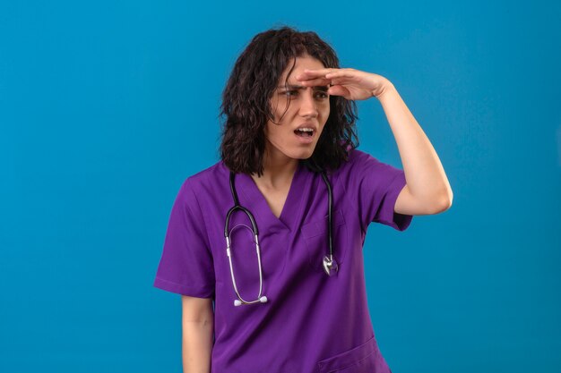 Mujer enfermera en uniforme médico y un estetoscopio mirando lejos con la mano para mirar algo de pie en azul aislado